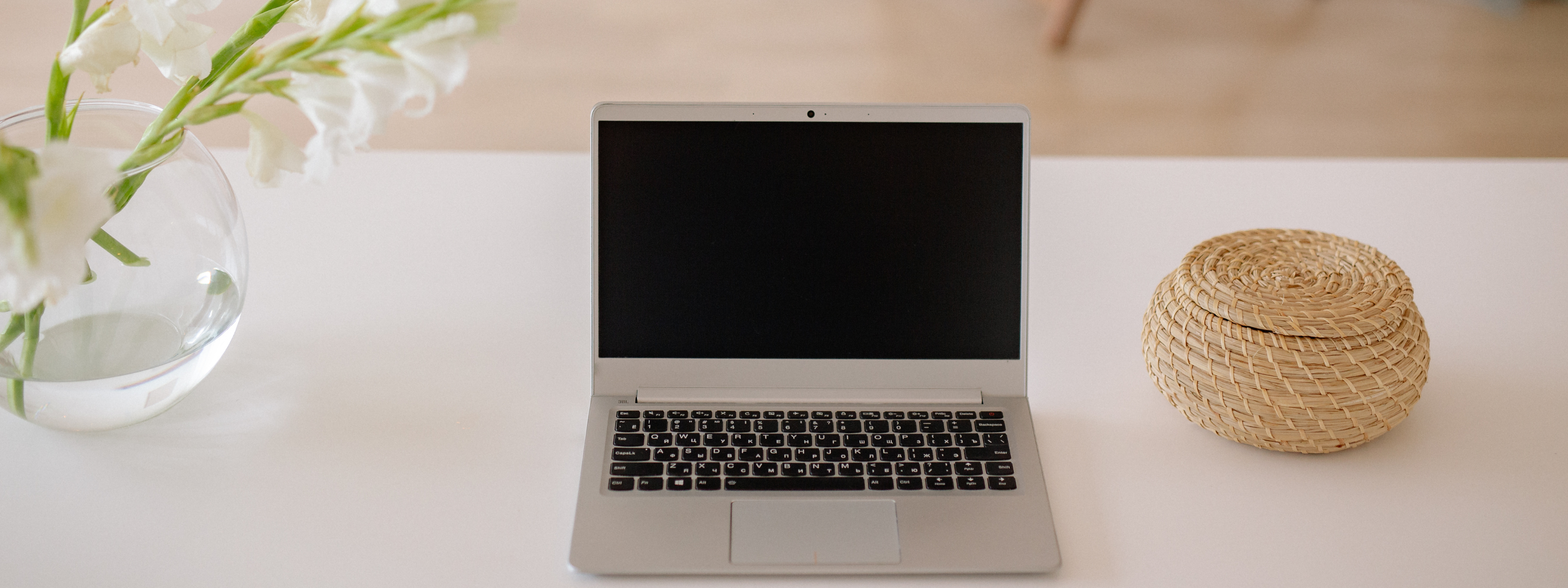 Laptop on a white surface with a vase of flowers and woven basket in the background