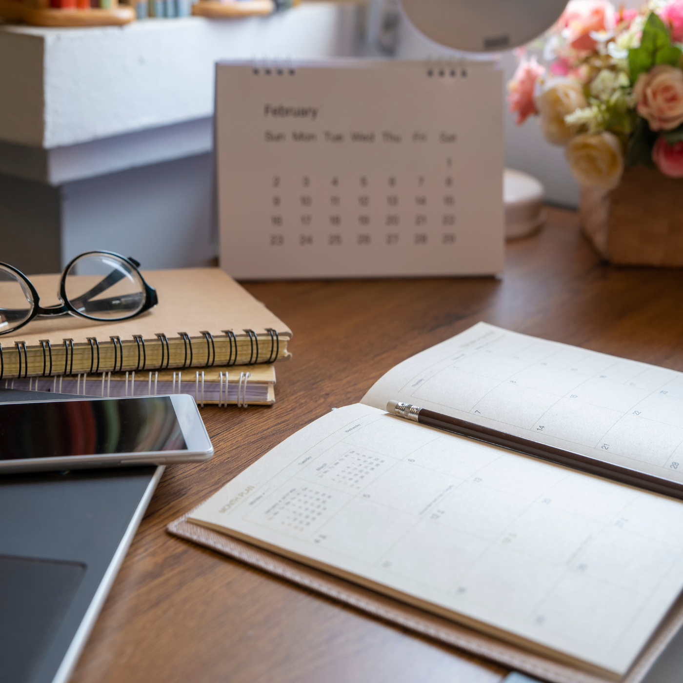 Desk with open notebook, pen, tablet, glasses, and calendar in a home office setting.