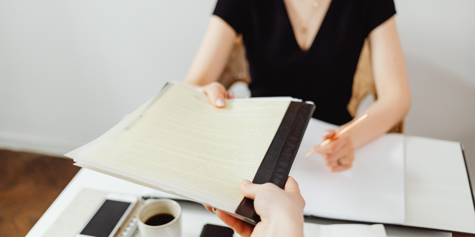 Person holding a folder with another person's hand reaching out to take it, on a desk with a coffee cup.