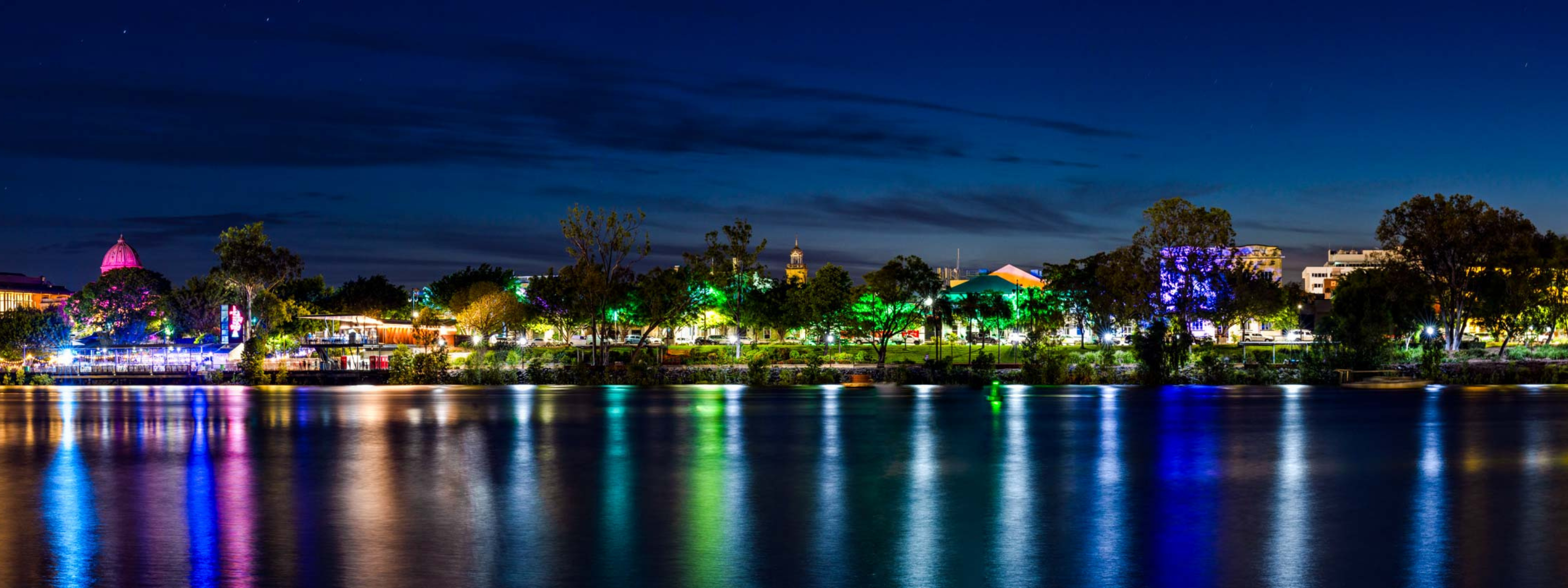 Panoramic view of a cityscape at night with illuminated buildings reflected in a body of water.